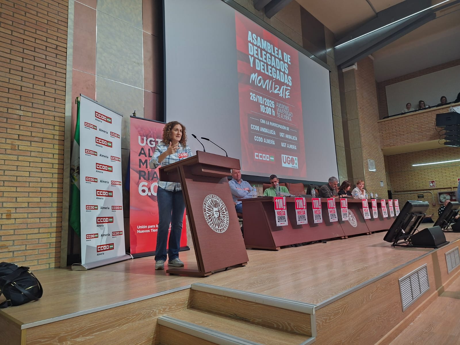 Una mujer habla en un atril en un escenario, ante una mesa con varias personas sentadas y una pantalla con el texto Asamblea de Delegados y Delegadas.