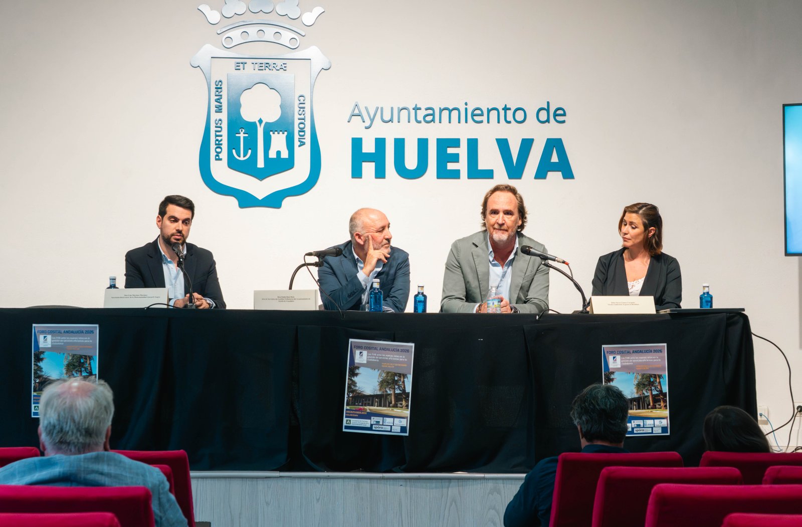 Cuatro personas sentadas tras una mesa con micrófonos, ante un panel con el escudo y el texto Ayuntamiento de Huelva, con público en butacas rojas.