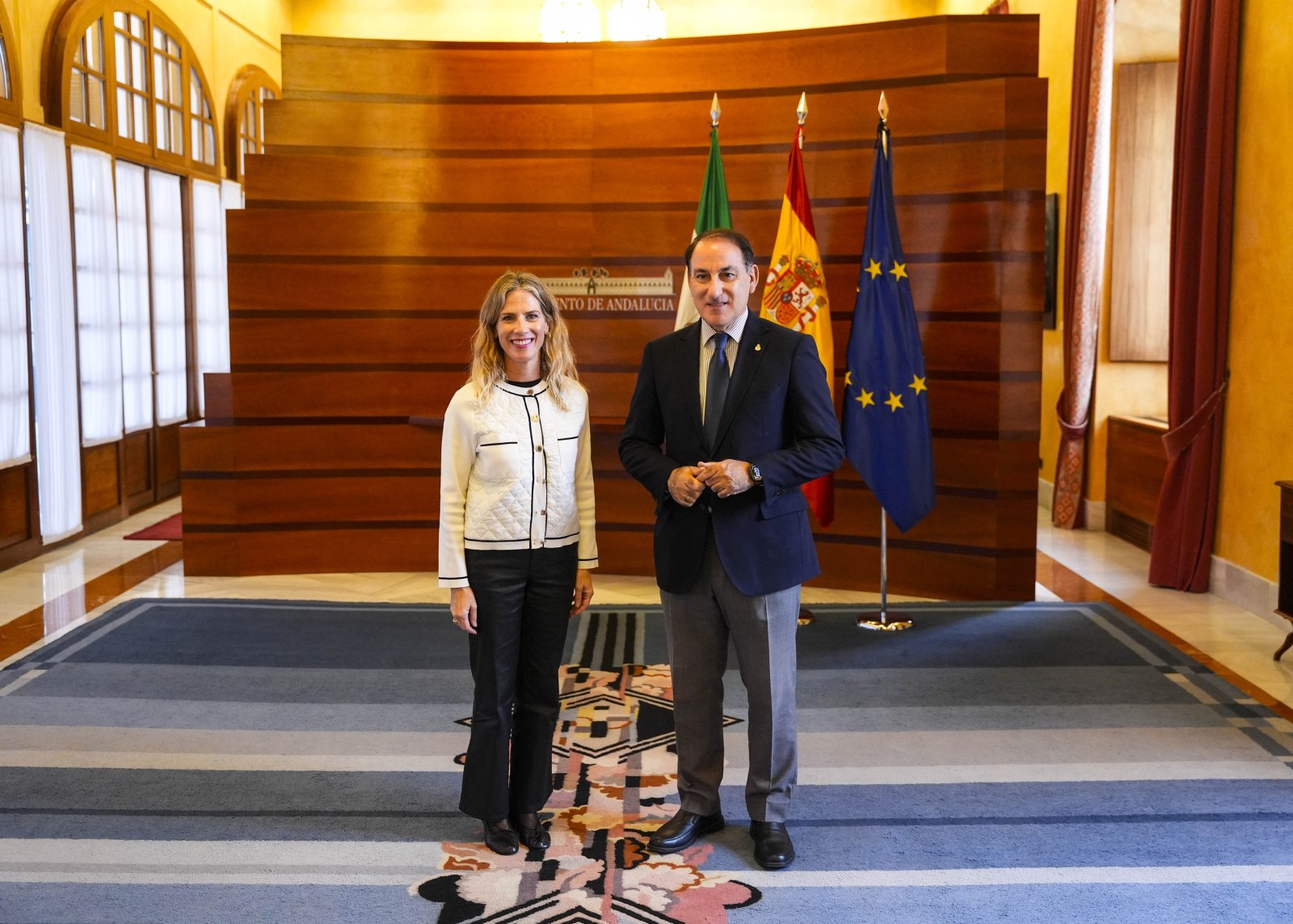 Un hombre con traje azul y una mujer con chaqueta blanca posan de pie en una sala con banderas de Andalucía, España y la UE.