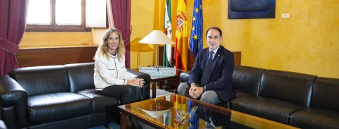 Un hombre con traje y una mujer con chaqueta blanca posan sentados en sofás de cuero frente a una mesa, con banderas al fondo.