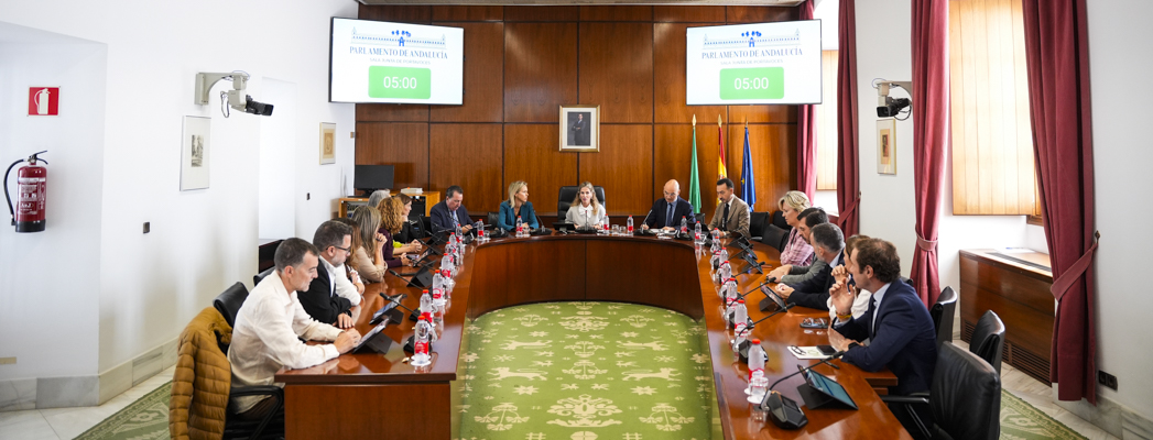 Personas sentadas alrededor de una mesa ovalada en una sala de reuniones con pantallas que marcan 05:00 y banderas al fondo.
