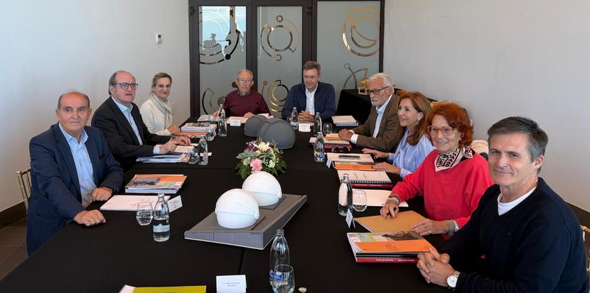 Varias personas sentadas alrededor de una mesa de reuniones alargada con carpetas, botellas y un centro floral, en una sala con puertas de cristal decoradas.