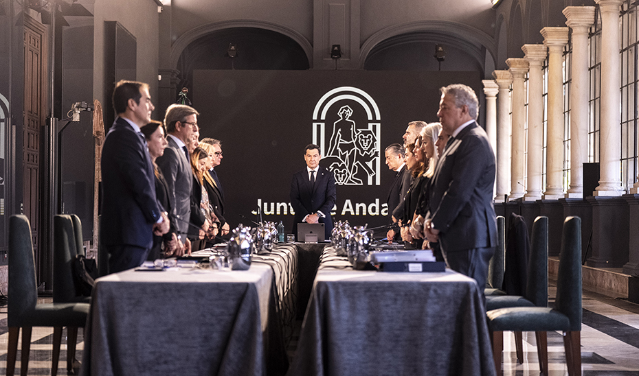 Varias personas trajeadas de pie junto a una mesa larga en una sala con columnas y un panel con un emblema y texto institucional.
