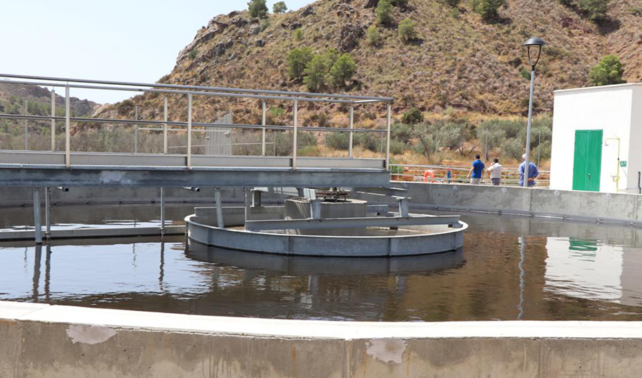 Depósitos circulares de agua con pasarela metálica, un edificio blanco con puerta verde y varias personas al fondo junto a una ladera.