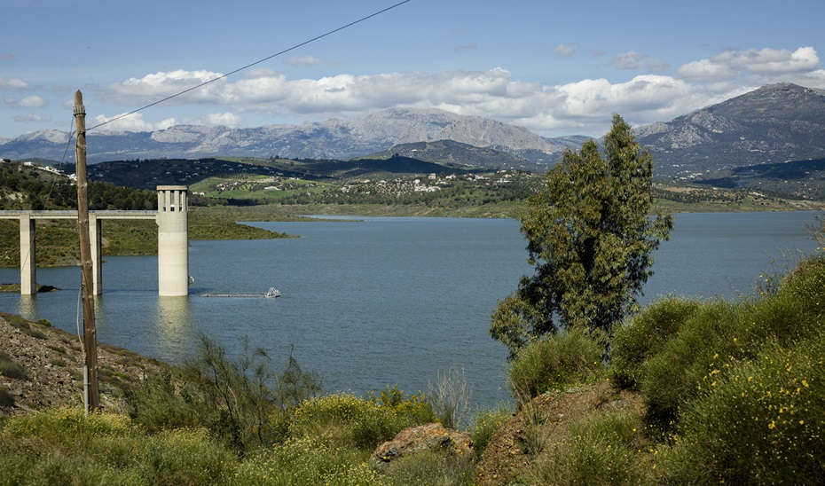 Un embalse con una torre de toma y un puente sobre el agua, rodeado de vegetación y montañas al fondo.
